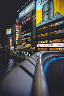 A vibrant urban scene featuring illuminated billboards on a building at night. The foreground shows a curved railing leading into the scene, with a focus on a bright advertisement for a beverage. The surrounding buildings have various lit signs and advertisements, contributing to the lively city atmosphere.