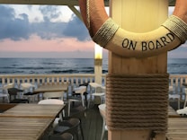 A serene beachfront scene with a wooden deck featuring several tables and chairs. A life preserver with 'ON BOARD' written on it is prominently displayed in the foreground. The ocean waves gently meet the shore under a sky with soft pink and gray clouds, creating a calming atmosphere.