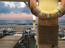 A serene beachfront scene with a wooden deck featuring several tables and chairs. A life preserver with 'ON BOARD' written on it is prominently displayed in the foreground. The ocean waves gently meet the shore under a sky with soft pink and gray clouds, creating a calming atmosphere.