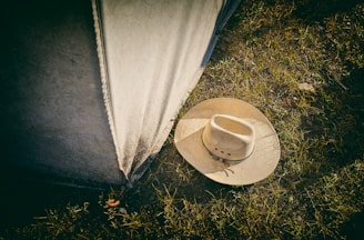 A beige cowboy hat lies on grassy ground next to a weathered canvas or tent wall. The grass is sparse, with dirt visible between the blades.