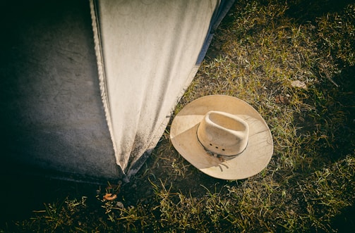 A beige cowboy hat lies on grassy ground next to a weathered canvas or tent wall. The grass is sparse, with dirt visible between the blades.