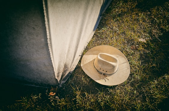 A beige cowboy hat lies on grassy ground next to a weathered canvas or tent wall. The grass is sparse, with dirt visible between the blades.