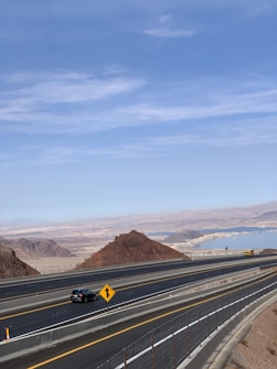 A highway stretches through a desert landscape with light brown rocky hills and mountains in the background. The road is divided into multiple lanes with vehicles visible, including a black car and a yellow truck. A road sign is present, and in the distance, a large body of water under a clear blue sky can be seen.