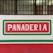 A sign with the word 'PANADERIA' in bold white letters on a red background surrounded by a green and white rectangular border, mounted on a wall with bars on the windows above.