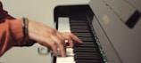 A close-up of a person's hand playing a piano. The fingers are poised over the black and white keys, with a blurred depth of field highlighting the hand and part of the piano.