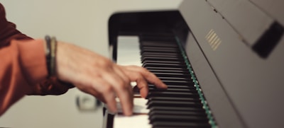 A close-up of a person's hand playing a piano. The fingers are poised over the black and white keys, with a blurred depth of field highlighting the hand and part of the piano.