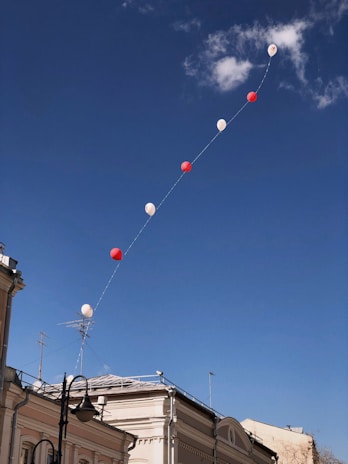 A series of red and white balloons are floating in a clear blue sky, tethered in a line. There are a few scattered clouds in the background. Below, the corner of a building with a classical architectural style is visible, featuring a rooftop and antennas.