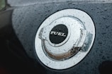 A close-up view of a car's metallic fuel cap covered in water droplets. The word 'FUEL' is prominently displayed on the cap. The surface has a shiny, reflective quality enhanced by the moisture.