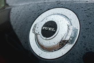 A close-up view of a car's metallic fuel cap covered in water droplets. The word 'FUEL' is prominently displayed on the cap. The surface has a shiny, reflective quality enhanced by the moisture.
