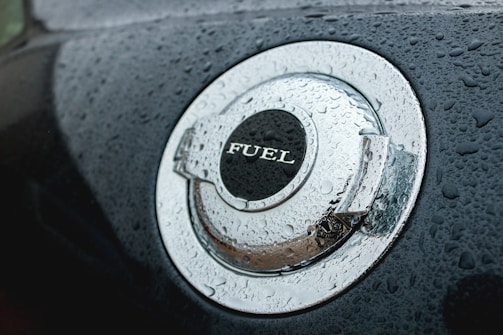 A close-up view of a car's metallic fuel cap covered in water droplets. The word 'FUEL' is prominently displayed on the cap. The surface has a shiny, reflective quality enhanced by the moisture.