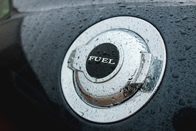 A close-up view of a car's metallic fuel cap covered in water droplets. The word 'FUEL' is prominently displayed on the cap. The surface has a shiny, reflective quality enhanced by the moisture.