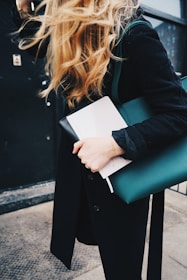 A person with long, wavy blond hair is holding a green leather bag and a book or notebook. They are dressed in a black coat and wearing a delicate bracelet on their wrist. The background includes a dark wall and a metal door, suggesting an urban setting.