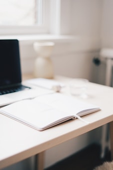 A clean, minimalist desk space with an open notebook in the center, a closed laptop on the left, a glass of water to the right, and a decorative vase in the background. The desk is light-colored, positioned next to a window allowing natural light to brighten the space.