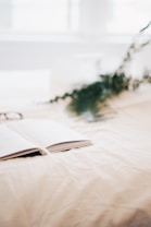 An open notebook lies on a soft, cream-colored surface with leafy green branches in the background. The overall composition is softly focused, creating a serene and inviting atmosphere.