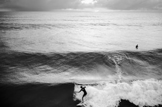 A black and white photograph capturing a surfer riding a wave in the foreground, with another surfer positioned further out in the ocean. The scene is set under a cloudy sky that enhances the dramatic atmosphere of the ocean.