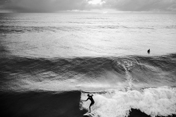 A black and white photograph capturing a surfer riding a wave in the foreground, with another surfer positioned further out in the ocean. The scene is set under a cloudy sky that enhances the dramatic atmosphere of the ocean.