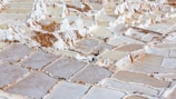 A vast expanse of terraced salt evaporation ponds, featuring a mosaic of rectangular pools with uneven stone borders. The ponds contain different levels of water, appearing in shades of white, beige, and light brown. The area is surrounded by natural rocky terrain.