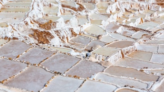 A vast expanse of terraced salt evaporation ponds, featuring a mosaic of rectangular pools with uneven stone borders. The ponds contain different levels of water, appearing in shades of white, beige, and light brown. The area is surrounded by natural rocky terrain.