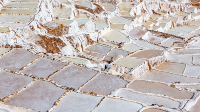 A vast expanse of terraced salt evaporation ponds, featuring a mosaic of rectangular pools with uneven stone borders. The ponds contain different levels of water, appearing in shades of white, beige, and light brown. The area is surrounded by natural rocky terrain.