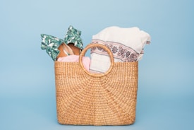 A woven straw basket is filled with various items, including a patterned piece of fabric with green leaves, sandals, and a white shirt with black and pink embroidery. The background is a solid light blue color, creating a serene and summer-like atmosphere.