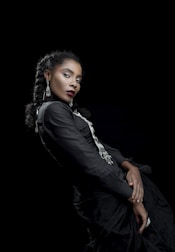 A woman with braided hair dressed in a black outfit with silver accents, posing against a dark background. Her expression is poised and confident, and she is adorned with long dangling earrings. The composition highlights elegance and sophistication.
