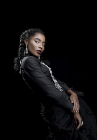 A woman with braided hair dressed in a black outfit with silver accents, posing against a dark background. Her expression is poised and confident, and she is adorned with long dangling earrings. The composition highlights elegance and sophistication.