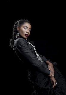 A woman with braided hair dressed in a black outfit with silver accents, posing against a dark background. Her expression is poised and confident, and she is adorned with long dangling earrings. The composition highlights elegance and sophistication.