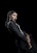 A woman with braided hair dressed in a black outfit with silver accents, posing against a dark background. Her expression is poised and confident, and she is adorned with long dangling earrings. The composition highlights elegance and sophistication.