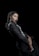 A woman with braided hair dressed in a black outfit with silver accents, posing against a dark background. Her expression is poised and confident, and she is adorned with long dangling earrings. The composition highlights elegance and sophistication.