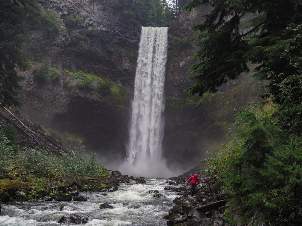 person wearing red jacket near waterfalls