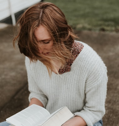 woman holding book while sitting on gray concrete pavement