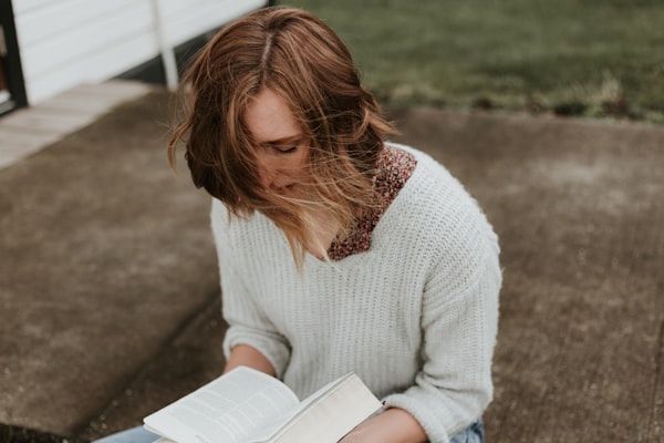 a woman in a white sweater, her hair ruffled by the wind, kneeling on a porch reading a book