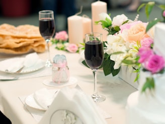 A table set for a formal event, featuring two glasses of red wine, floral arrangements with roses and white flowers, beige candles, and neatly folded napkins. There are also plates with utensils and a jar with a decorative ribbon.