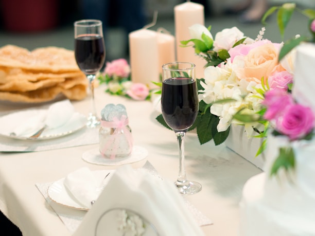 A table set for a formal event, featuring two glasses of red wine, floral arrangements with roses and white flowers, beige candles, and neatly folded napkins. There are also plates with utensils and a jar with a decorative ribbon.