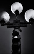 A black and white image featuring a traffic light and street lamp combination. Three spherical lights are positioned at the top, casting a subtle glow, while the traffic light is deactivated, its bulbs appearing dark. The background is a gradient of dark grey and black, emphasizing the silhouette of the structure.