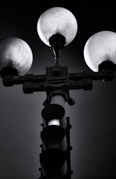 A black and white image featuring a traffic light and street lamp combination. Three spherical lights are positioned at the top, casting a subtle glow, while the traffic light is deactivated, its bulbs appearing dark. The background is a gradient of dark grey and black, emphasizing the silhouette of the structure.