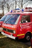 A red fire service vehicle with the words 'FEUERWEHR' and 'Jugendfeuerwehr Mindelheim' painted on the side. The vehicle has a Volkswagen emblem on the front. It is parked outdoors on grass with other vehicles nearby. There are emergency lights and loudspeakers mounted on the roof.