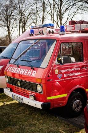A red fire service vehicle with the words 'FEUERWEHR' and 'Jugendfeuerwehr Mindelheim' painted on the side. The vehicle has a Volkswagen emblem on the front. It is parked outdoors on grass with other vehicles nearby. There are emergency lights and loudspeakers mounted on the roof.