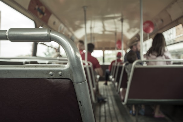 A vintage-style bus interior with metal handrails and wooden floorboards. Several passengers, including adults and a child, are seated and facing forward. The atmosphere inside is calm, with muted lighting and slightly worn upholstery.