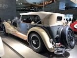 A classic vintage car with a beige soft top and shiny chrome accents is on display indoors. The body is mainly a sleek gray color with polished wooden running boards. A spare tire is mounted at the rear alongside detailed ornate features. Overhead, a vintage aircraft suspended from the ceiling adds to the retro ambiance.