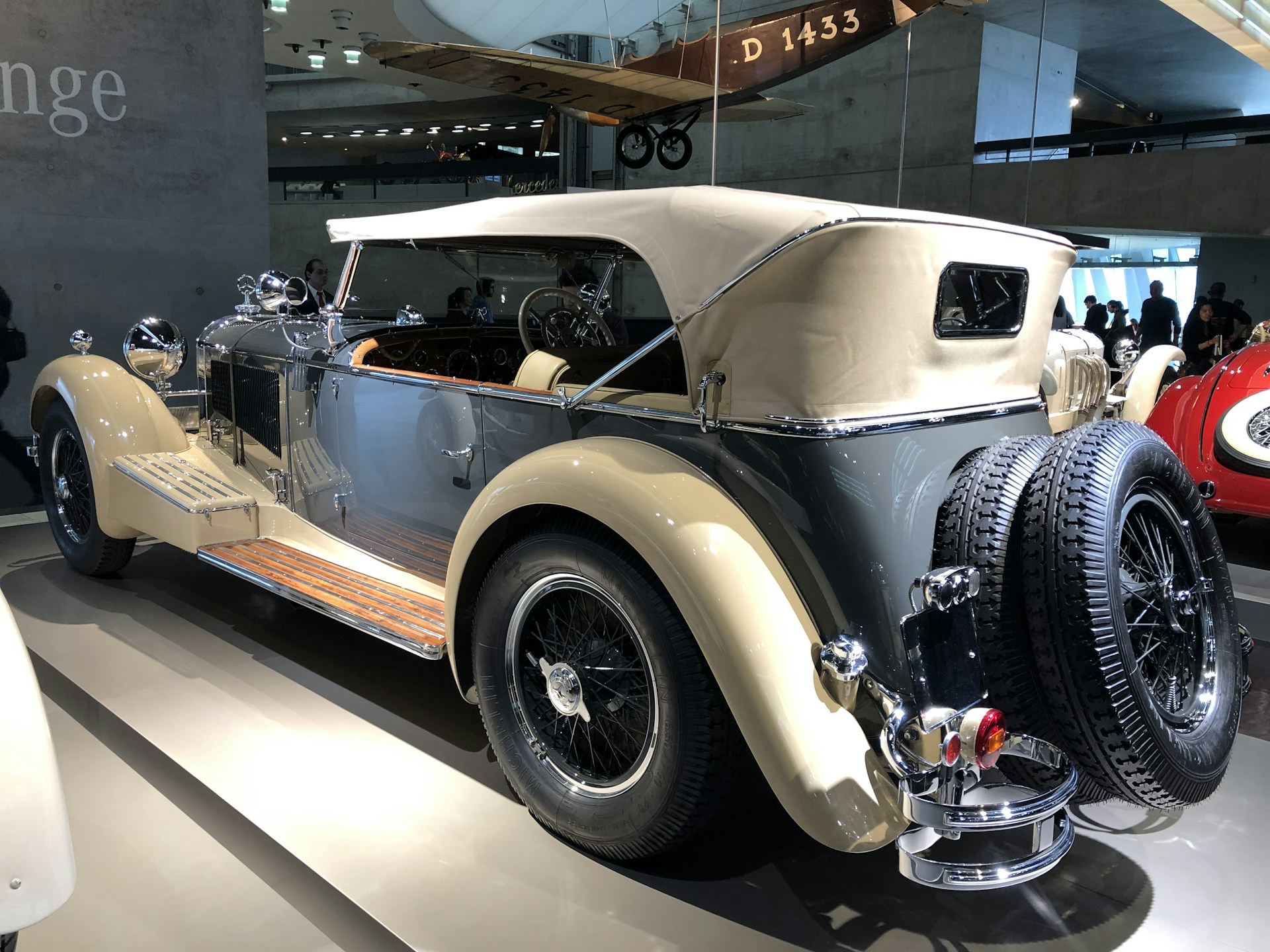 A classic vintage car with a beige soft top and shiny chrome accents is on display indoors. The body is mainly a sleek gray color with polished wooden running boards. A spare tire is mounted at the rear alongside detailed ornate features. Overhead, a vintage aircraft suspended from the ceiling adds to the retro ambiance.