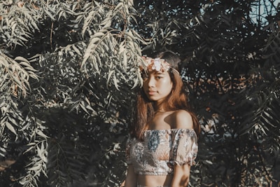 A young woman stands amidst lush foliage, with sunlight casting dappled shadows on her. She wears an off-the-shoulder floral top and a headband adorned with pink flowers, contributing to a bohemian aesthetic.