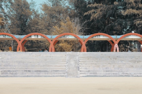 A series of red brick arches create an architectural feature in front of a backdrop of dense, tall trees. The structure stands on a stepped concrete platform with an open sandy ground in the foreground. A few people are visible beneath the arches, suggesting it is a public or recreational space.