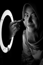 A black-and-white image of a person wearing a headscarf, smiling, and holding a makeup brush near their cheek. There is a round light source illuminating the face from the left side.