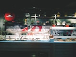 A salad bar is displayed with various pans of ingredients under a glass cover. Behind the counter are shelves with plants, pots, and jars. The wall behind features graphic designs with the text 'Making Awesome Pizza' and a large '2 Live Lyfe'. A red ceiling light fixture is visible on the upper left.