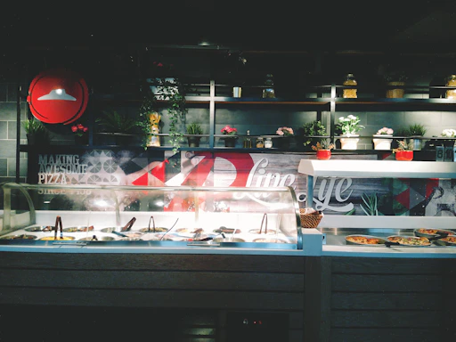 A vibrant display of fresh salads, sandwiches, and pizzas on a rustic wooden table inside a bright, open kitchen.