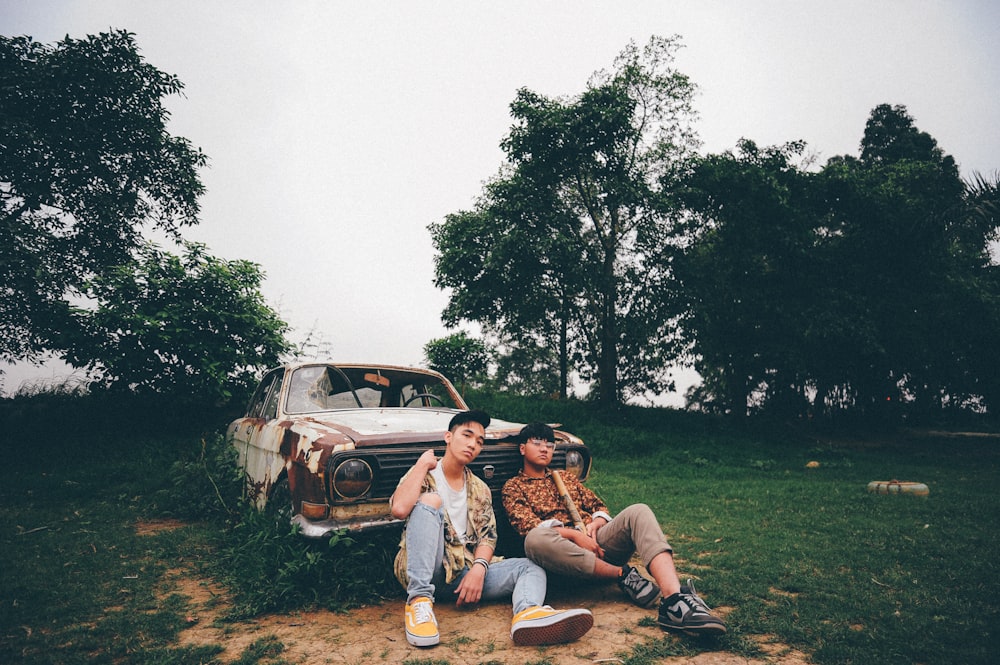 two men sitting on ground while leaning on abandoned white vehicle during daytime