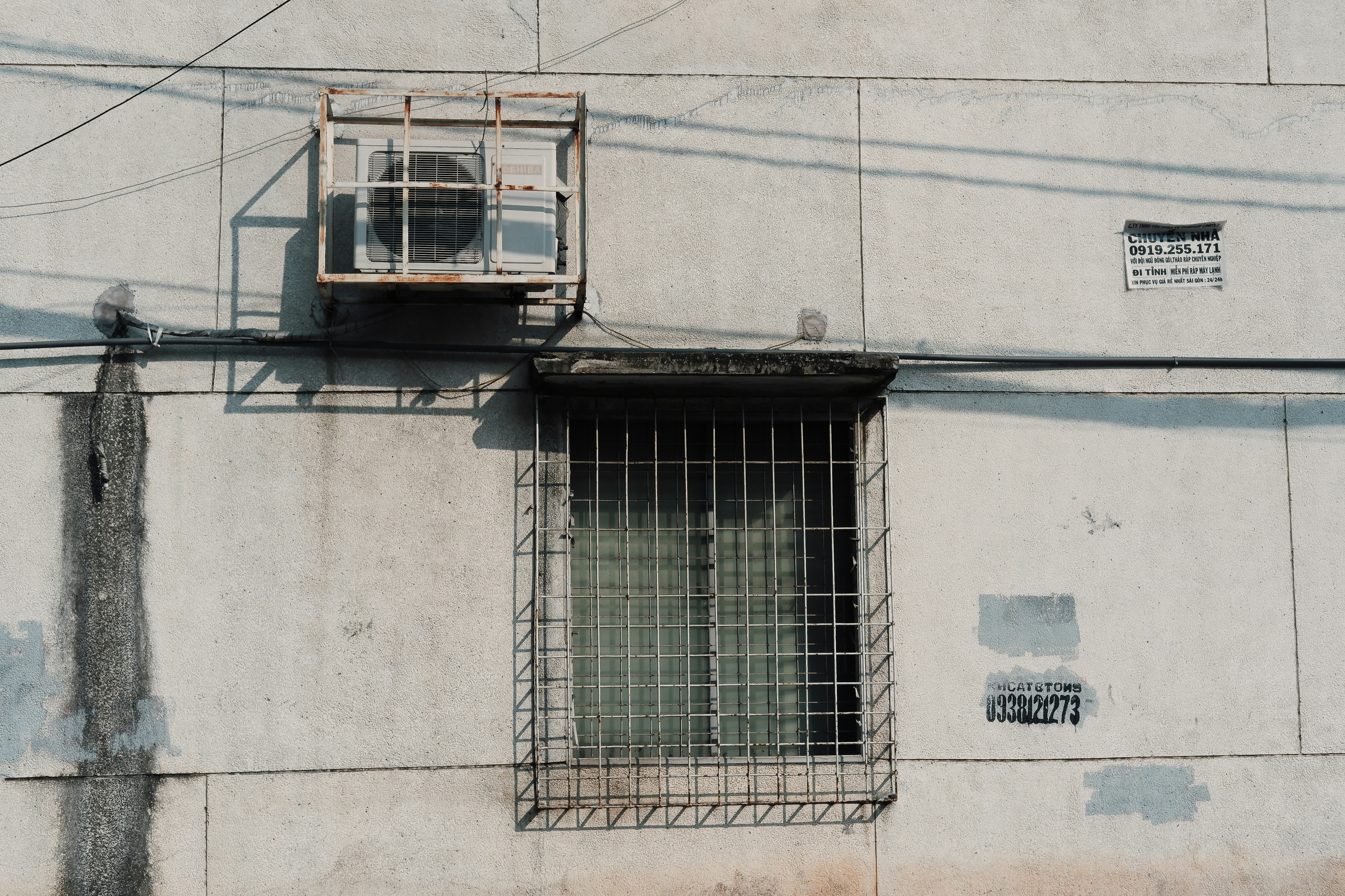 A concrete wall with a rusted air conditioning unit mounted above a barred window. The wall displays faded paint patches and two stenciled phone numbers. Overhead electrical wires cast shadows across the surface.