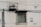 A concrete wall with a rusted air conditioning unit mounted above a barred window. The wall displays faded paint patches and two stenciled phone numbers. Overhead electrical wires cast shadows across the surface.