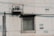 A concrete wall with a rusted air conditioning unit mounted above a barred window. The wall displays faded paint patches and two stenciled phone numbers. Overhead electrical wires cast shadows across the surface.